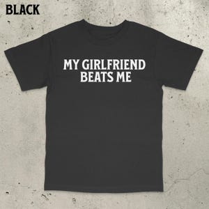 May include: Black t-shirt with the text "MY GIRLFRIEND BEATS ME" in white block letters. The shirt is a crew neck style and appears to be made of a soft material. The word "BLACK" is in the top left corner.