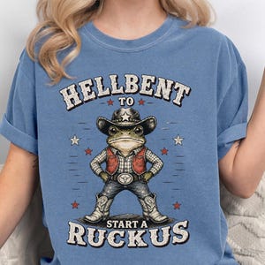 May include: A blue short-sleeve t-shirt featuring a graphic of a frog wearing a cowboy hat, vest, and boots. The text reads HELLBENT TO START A RUCKUS. The design includes red and white stars surrounding the frog, with a distressed, vintage-style print.
