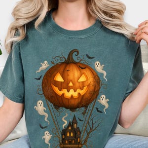 May include: Teal-colored t-shirt featuring a Halloween-themed design. The graphic includes a jack-o'-lantern with a smiling face, surrounded by ghosts and bats. Below the pumpkin is a haunted house in a basket, with bare tree branches.