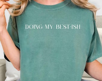 May include: A teal-colored t-shirt with the phrase "DOING MY BEST-ISH" printed in white capital letters. The shirt has a crew neck and short sleeves. The fabric appears soft and slightly faded.