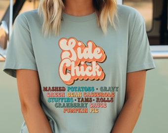 May include: A light blue t-shirt with the text "Side Chick" in a retro font. The text is in orange, pink, and yellow. Below the text is a list of Thanksgiving side dishes in a variety of colors: mashed potatoes, gravy, green bean casserole, stuffing, yams, rolls, cranberry sauce, and pumpkin pie.