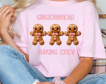 May include: A light pink t-shirt featuring the text "GINGERBREAD BAKING CREW" above three gingerbread man graphics. The gingerbread men have brown bodies, pink bow ties, and decorative icing.