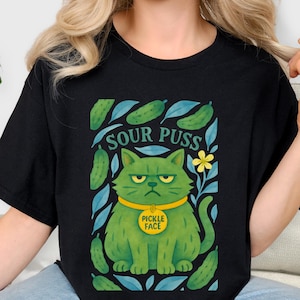 May include: Black t-shirt featuring a green cat illustration with the text "SOUR PUSS" above it. The cat has a yellow collar that says "PICKLE FACE". The design includes pickles and blue leaves.
