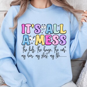 May include: Light blue sweatshirt with the text "IT'S ALL A MESS" in colorful bubble letters. The text below lists "the kids, the house, the car, my hair, my shirt, my life..." in a black script font. The sweatshirt is a casual style.