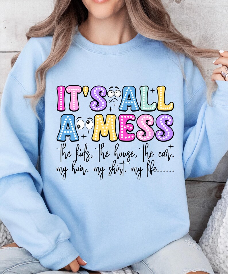 May include: Light blue sweatshirt with the text "IT'S ALL A MESS" in colorful bubble letters. The text below lists "the kids, the house, the car, my hair, my shirt, my life..." in a black script font. The sweatshirt is a casual style.