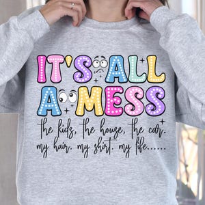 May include: A light gray sweatshirt with the text "IT'S ALL A MESS" in colorful bubble letters. Below the text are the words "the kids, the house, the car. my hair. my shirt. my life....." in a handwritten font.