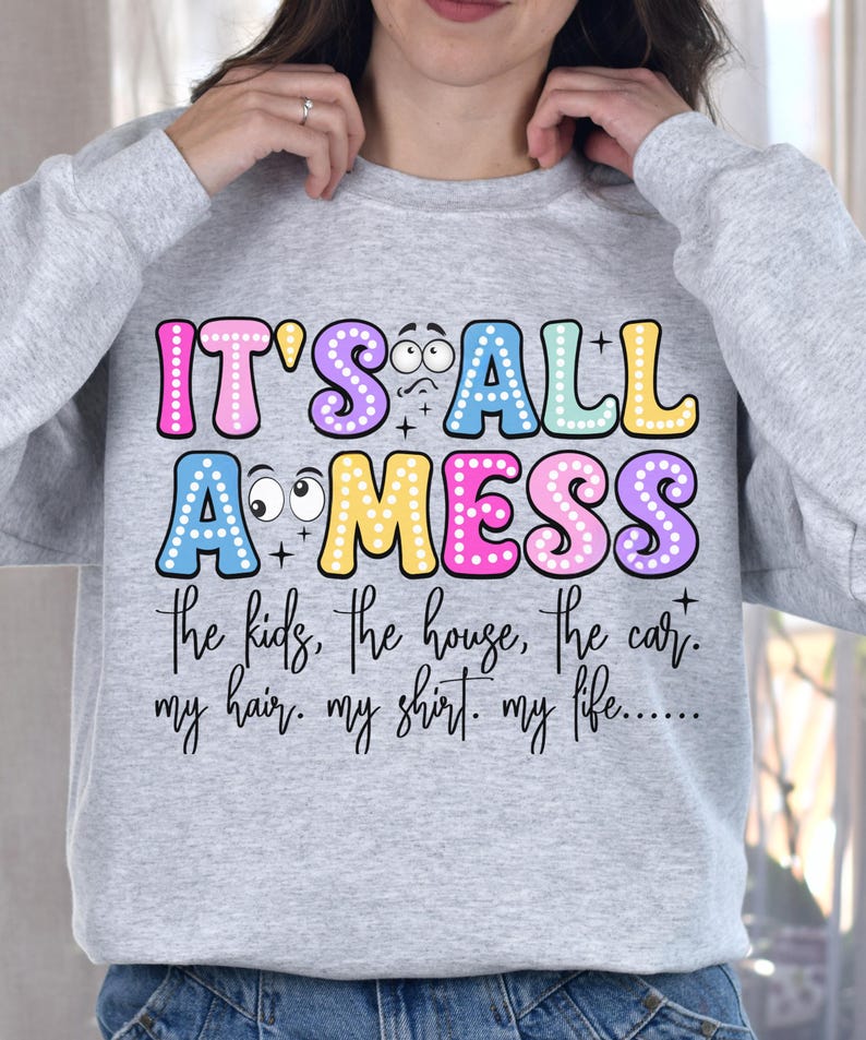 May include: A light gray sweatshirt with the text "IT'S ALL A MESS" in colorful bubble letters. Below the text are the words "the kids, the house, the car. my hair. my shirt. my life....." in a handwritten font.