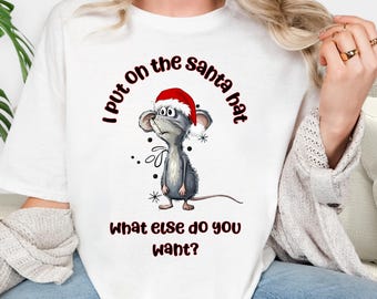May include: White t-shirt featuring a cartoon mouse wearing a Santa hat. The text reads "I put on the santa hat" and "What else do you want?" in a festive font. The design includes snowflakes and dots.