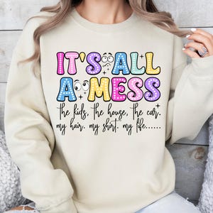 May include: A beige sweatshirt with the text "IT'S ALL A MESS" in colorful bubble letters. The text below reads "the kids, the house, the car. my hair. my shirt. my life....." A casual, humorous design.