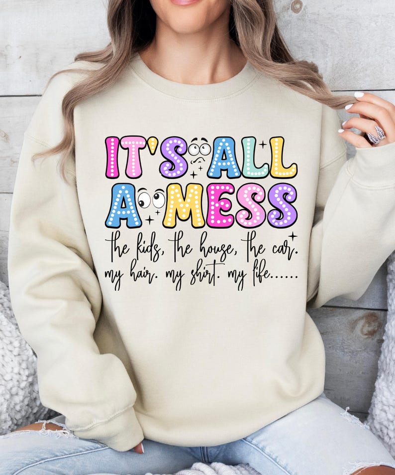 May include: A beige sweatshirt with the text "IT'S ALL A MESS" in colorful bubble letters. The text below reads "the kids, the house, the car. my hair. my shirt. my life....." A casual, humorous design.