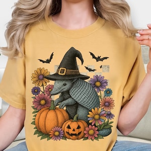May include: Mustard yellow t-shirt featuring a Halloween-themed illustration. The design includes an armadillo wearing a witch hat, pumpkins, flowers, and bats. The graphic is colorful and whimsical, perfect for the spooky season.