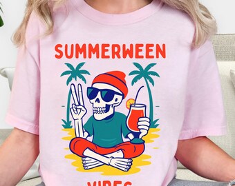 May include: Pink t-shirt with the words "SUMMERWEEN VIBES" in red and white. The graphic features a skeleton wearing sunglasses and a beanie, holding a cocktail and making a peace sign. Palm trees are in the background.