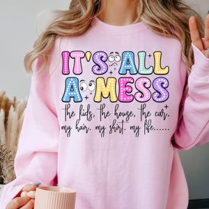 May include: A light pink sweatshirt with the text "IT'S ALL A MESS" in colorful bubble letters. The text below reads "the kids, the house, the car. my hair. my shirt. my life..." The sweatshirt is a casual style.