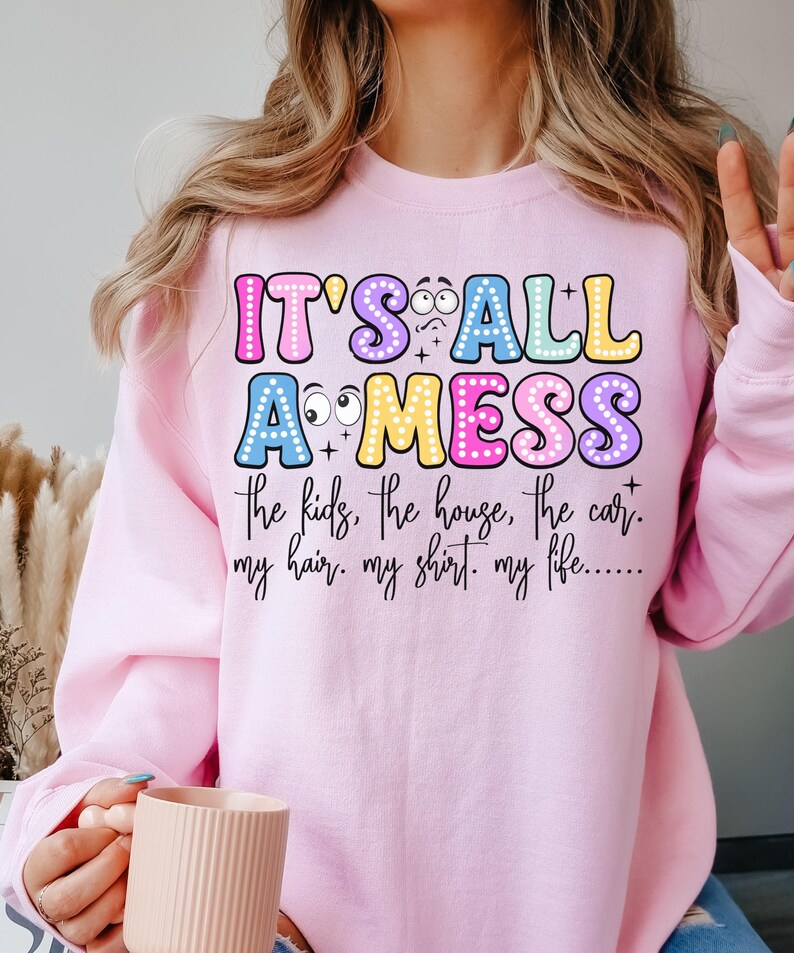 May include: A light pink sweatshirt with the text "IT'S ALL A MESS" in colorful bubble letters. The text below reads "the kids, the house, the car. my hair. my shirt. my life..." The sweatshirt is a casual style.