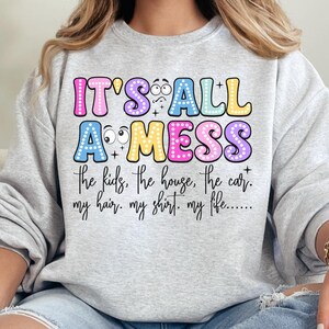 May include: A light gray sweatshirt with the text "IT'S ALL A MESS" in colorful bubble letters. Below the text are the words "the kids, the house, the car. my hair. my shirt. my life....."