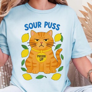 May include: Light blue t-shirt with the text "SOUR PUSS" above a cartoon orange cat with a grumpy expression. The cat wears a yellow collar that says "PUCKER UP". Lemons and green leaves surround the cat.
