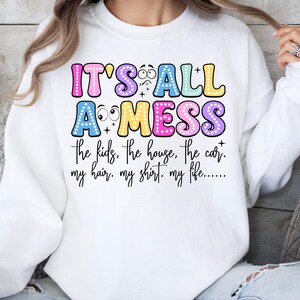 May include: White sweatshirt with the text "IT'S ALL A MESS" in colorful bubble letters. The text continues with "the kids, the house, the car, my hair, my shirt, my life..." in a handwritten style font. The sweatshirt has long sleeves.