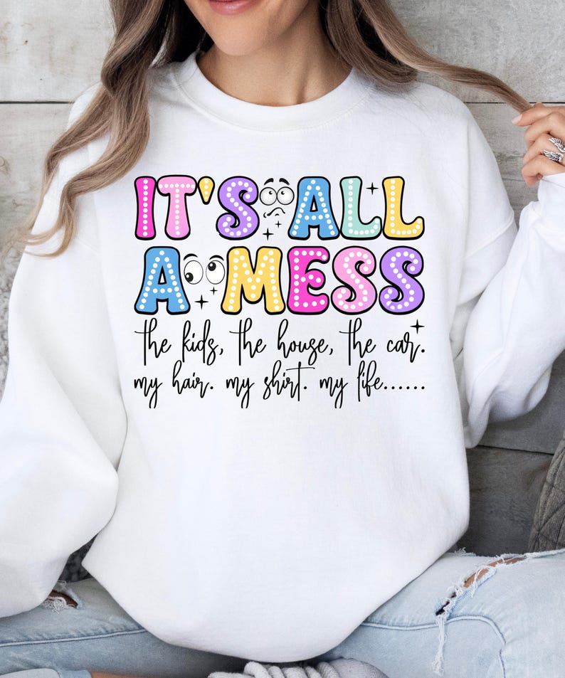 May include: White sweatshirt with the text "IT'S ALL A MESS" in colorful bubble letters. The text continues with "the kids, the house, the car, my hair, my shirt, my life..." in a handwritten style font. The sweatshirt has long sleeves.