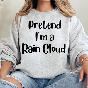 May include: A light gray sweatshirt with the text "Pretend I'm a Rain Cloud" in black. The sweatshirt is a crew neck style and is made of a soft material. The image shows a person wearing the sweatshirt.