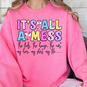 May include: A bright pink sweatshirt with the text "IT'S ALL A MESS" in colorful bubble letters. Below the text are the words "the kids, the house, the car. my hair. my shirt. my life......"