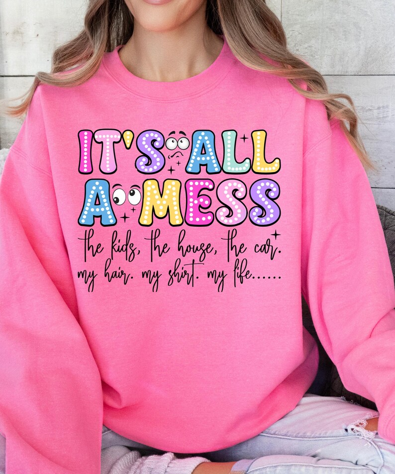 May include: A bright pink sweatshirt with the text "IT'S ALL A MESS" in colorful bubble letters. Below the text are the words "the kids, the house, the car. my hair. my shirt. my life......"