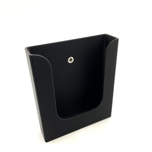 May include: Black plastic wall mount with a single screw visible. The mount has a rectangular opening for holding an object.