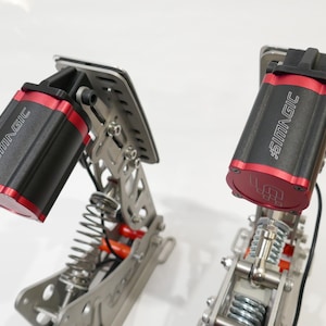 Puede incluir: Dos pedales de simulador de carreras negros y rojos con un mecanismo de resorte y el texto "#SIMMAGIC" en el lateral.