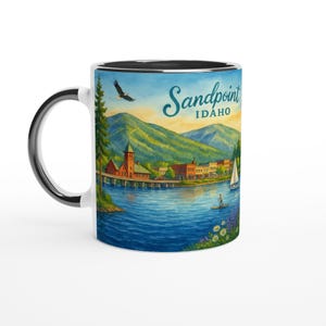 Op de afbeelding: Witte keramische mok met een schilderachtige aquarel illustratie van Sandpoint, Idaho. Het ontwerp omvat een meer, bergen, gebouwen en de tekst "Sandpoint Idaho". De mok heeft een zwarte binnenkant en handvat.