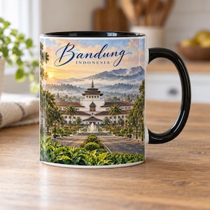 May include: A ceramic mug with a black handle and interior, featuring a printed landscape image of the Gedung Sate building in Bandung, Indonesia. The text Bandung Indonesia is printed in blue script and block lettering above the building.