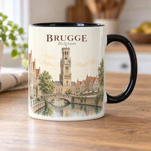 Op de afbeelding: Een witte keramische mok met een zwarte handgreep en zwarte binnenrand, voorzien van een gedetailleerde illustratie van het Belfort van Brugge, een stenen brug en gebouwen langs het kanaal. De tekst BRUGGE Belgium staat in bruin boven de toren.