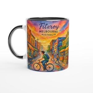May include: White ceramic mug with a black interior and handle, featuring a colorful watercolor illustration of Fitzroy, Melbourne, Australia. The design includes a person riding a bicycle on a street with shops and buildings, and the text "Fitzroy Melbourne Australia".