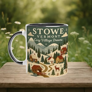 May include: White ceramic mug with a black interior and handle, featuring a scenic illustration of Stowe, Vermont. The design includes houses, trees, mountains, and the text "STOWE VERMONT Cozy Village Dreams". The mug sits on a wooden surface.