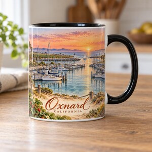 May include: A ceramic mug with a black handle and rim featuring a colorful illustration of a harbor at sunset with sailboats, a lighthouse, and the text Oxnard CALIFORNIA printed in a script font above the word CALIFORNIA.