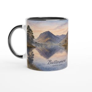 May include: White ceramic mug featuring a watercolor painting of a lake and mountains under a sunset sky. The mug has a black rim and handle, with the text "Buttermere ENGLAND" on the front.