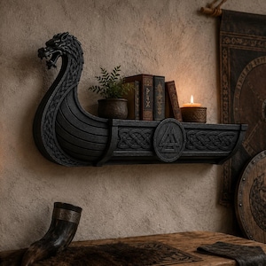 May include: A black wall-mounted shelf shaped like a Viking longship, featuring a carved dragon head prow and intricate Celtic knotwork patterns along the side. A circular emblem with a Valknut symbol is centred on the front of the shelf structure.