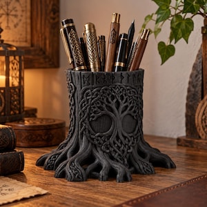May include: A black cylindrical pen holder shaped like a tree stump with exposed roots at the base. The exterior features a relief carving of a tree with intricate interwoven branches and a band of runic symbols around the top rim.