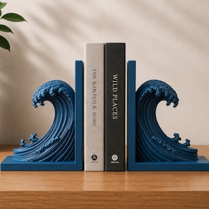 Puede incluir: Dos sujetalibros azules con forma de ola sostienen dos libros. Los sujetalibros son de color azul oscuro y presentan un diseño detallado de olas. Los libros se titulan "The Kinfolk Home" y "Wild Places". Están sobre una superficie de madera.