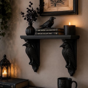 May include: A black wall-mounted shelf supported by two carved raven brackets. The shelf holds a black vase with dark foliage, a raven figurine, two books titled The Raven and Other Poems by Edgar Allan Poe, and a lit black pillar candle.