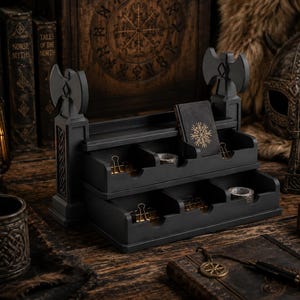May include: A dark grey desk tidy with two drawers and axe-shaped accents. It contains paper clips, tape, and a notebook with a gold snowflake design. The background features books, a lantern, and a Viking helmet, creating a themed aesthetic.