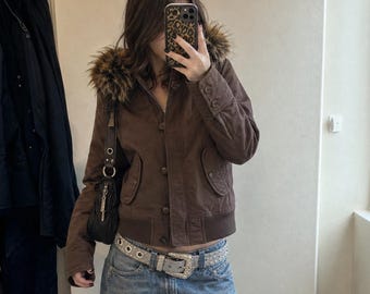 Peut inclure: Une veste marron avec une capuche bordée de fausse fourrure, une fermeture à boutons sur le devant et un ourlet côtelé. La veste est associée à un jean bleu et une ceinture argentée avec une boucle décorative et des œillets métalliques.