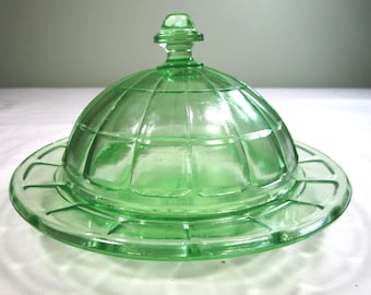 May include: A translucent light green glass butter dish with a domed lid. The lid features a grid pattern of raised squares and a central knob handle. The base has a matching grid pattern on the rim, creating a cohesive, geometric design.