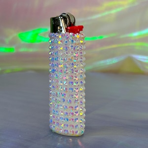May include: A white lighter covered in iridescent rhinestones. The lighter has a silver top and a red button.