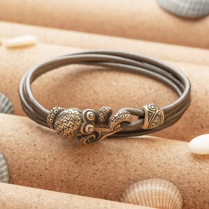 May include: A brown leather bracelet with a detailed octopus clasp. The bracelet is made of three strands of leather and has a decorative metal clasp.