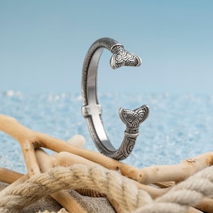 Könnte beinhalten: Ein silberfarbenes Armreif-Armband mit einem Meerjungfrauenschwanz-Design. Das Armband hat aufwendige Details und ist oben und unten offen. Der Hintergrund ist hellblau, mit Treibholz und Seil im Vordergrund.