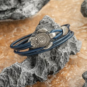 May include: A blue leather bracelet with a silver turtle charm. The bracelet is wrapped around a gray rock, showcasing the intricate details of the turtle design. The bracelet's leather strands are a deep blue color.