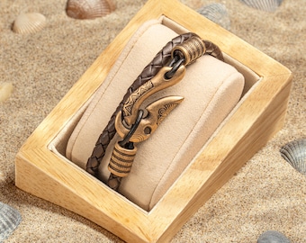 May include: A brown leather bracelet with a bronze-colored hook clasp, displayed in a wooden jewelry box with a beige interior. The bracelet has a braided design. The text "THE JEWELRY BOX IS NOT FOR SALE" is visible.