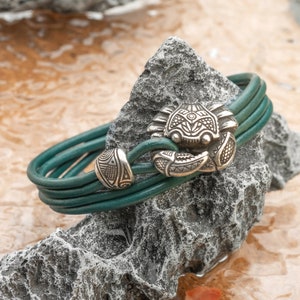 May include: A green leather bracelet with a silver crab charm. The bracelet is wrapped around a gray rock.