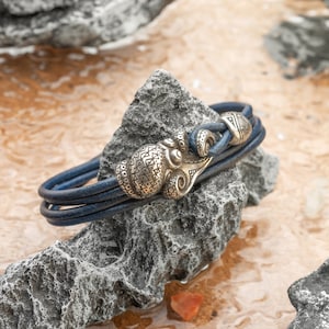 May include: A blue leather bracelet featuring an octopus-shaped clasp. The clasp is made of a metallic material with detailed patterns. The bracelet is displayed on a grey rock, highlighting the design and texture of the jewellery.