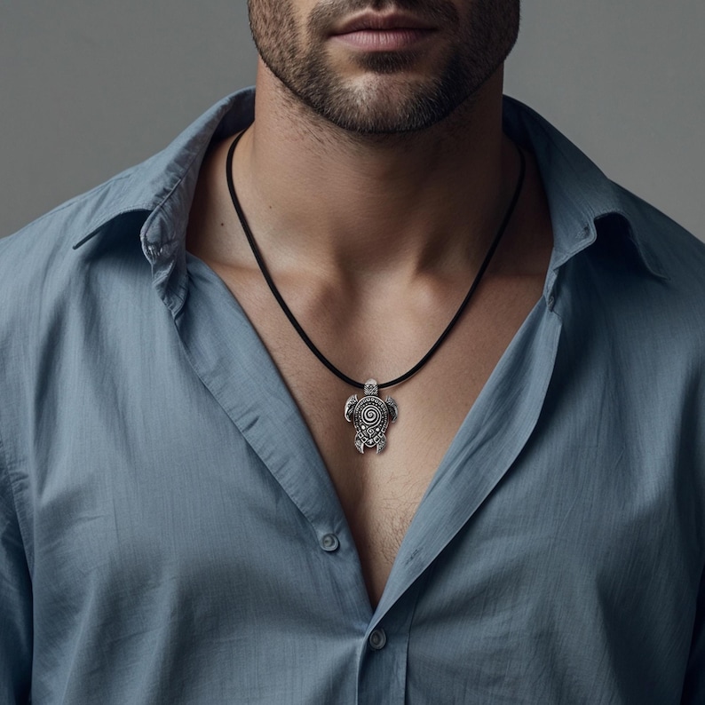 May include: A silver turtle pendant necklace with a black cord. The turtle pendant has detailed spiral and circular patterns. The necklace is shown against a blue button-down shirt.