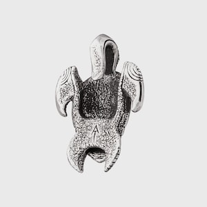 May include: A silver pendant shaped like a turtle with textured detailing. The pendant features wing-like elements and a curved head. A small letter "A" is engraved on the bottom. The pendant is likely made of a metal alloy.